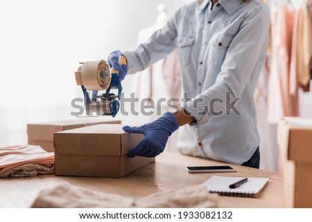 cropped view of seller in latex gloves sealing carton box in showroom, blurred foreground