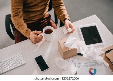 Cropped View Of Sick Businessman Holding Cup Of Tea And Box Of Tissues At Office Desk