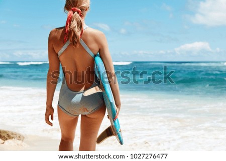 Cropped view of slim female model fond of surfing, stands back to ocean with surf board, admires blue clear sky, has fit bottom, wants some activity after lying on beach. People, summer sport concept