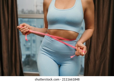 Cropped view of slim woman measuring waist with tape measure at home, close up. Unrecognizable european woman checking the result of diet for weight loss or liposuction indoors