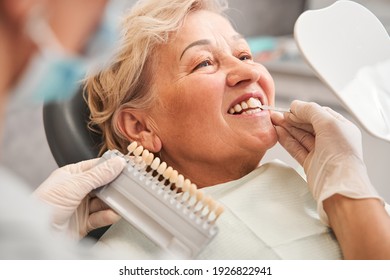 Cropped view of the smiling blonde woman patient having appointment in dental clinic while picking up shade using tooth enamel scale held by dentist in white gloves at dental clinic - Powered by Shutterstock