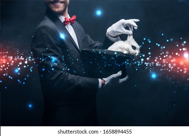 cropped view of smiling magician showing trick with white rabbit in hat, in dark room with smoke and glowing illustration