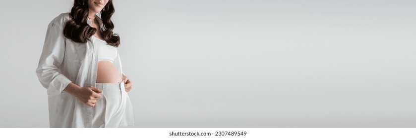 cropped view of smiling and pregnant woman in white fashionable shirt and pants standing isolated on grey background, maternity fashion concept, banner - Shutterstock ID 2307489549
