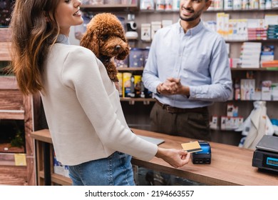 Cropped view of smiling woman holding poodle and paying with credit card near muslim seller in pet shop 