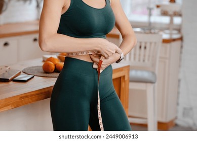 Cropped view of the sporty woman measuring waist with tape at bright kitchen. Weight loss and diet concept 