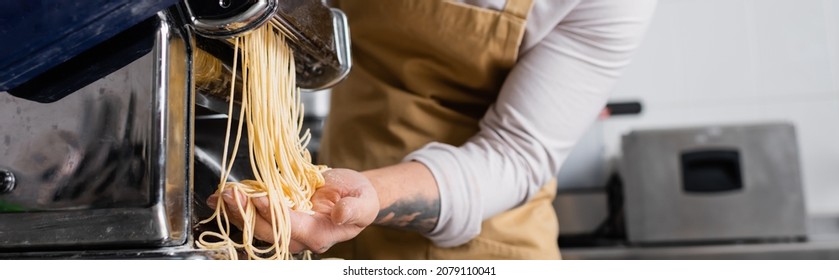 Cropped view of tattooed chef making spaghetti on pasta maker machine in kitchen, banner - Powered by Shutterstock