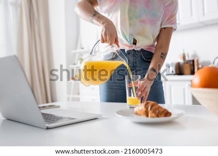 cropped view of tattooed woman pouring orange juice near croissant and blurred laptop