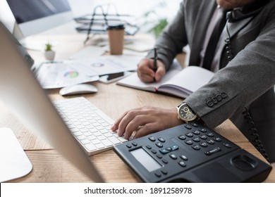 cropped view of trader talking on landline phone, writing in notebook and typing on laptop, blurred foreground - Shutterstock ID 1891258978