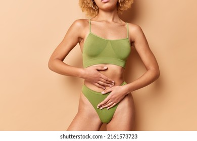 Cropped view of unrecognizable fit woman wears green top and panties keeps hands on flat belly poses against brown background shows perfect body after diet. Empty space for your advertisement