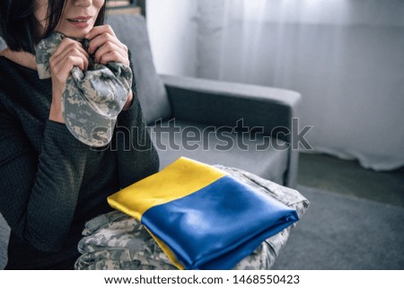 Cropped view of upset woman holding military clothing and ukrainian flag at home