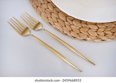 Cropped view of a white plate set on a woven rattan table mat with golden cutlery aside, minimalistic table arrangement in a restaurant, sitting area prepared for a special dinner or a formal event - Powered by Shutterstock