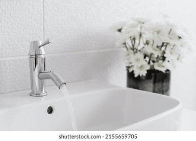 Cropped View Of White Sink With Flowing Water And Plants In Bathroom In White Tones. Scandinavian Room Interior Design. Home Comfort. Resource Saving. Home Comfort