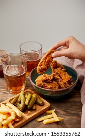 Cropped View Of Woman Eating Delicious Chicken Nuggets, French Fries And Gherkins Near Glasses Of Beer On Wooden Table Isolated On Grey