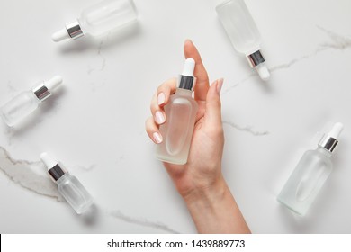 Cropped View Of Woman Hand With Moisturizer Serum Near Cosmetic Glass Bottles On White Surface