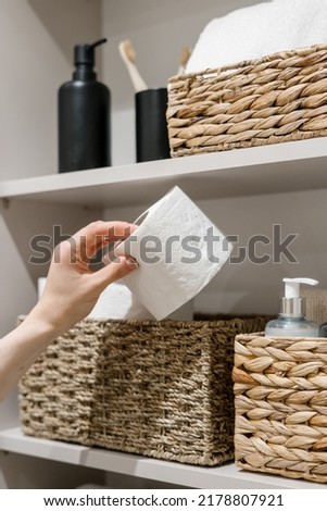 Cropped view of woman hand take toilet paper from wicker box. Domestic and household items on storage shelf in bathroom. Cosmetics bottles, clean and fresh towel in restroom closet