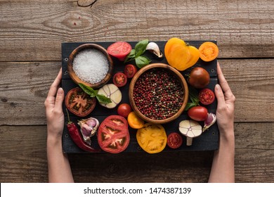 cropped view of woman holding black tray with pepper and salt in bowls, chilli pepper, sliced tomatoes and garlic on wooden table  - Powered by Shutterstock