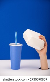 cropped view of woman holding carton box near paper cup with soft drink isolated on blue