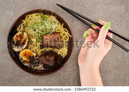 cropped view of woman holding chopsticks near ramen with egg and beef in bowl on grey background