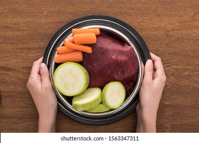 Cropped View Of Woman Holding Pet Bowl With Raw Meat And Vegetables On Wooden Background