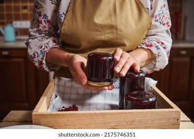 Cropped view of a woman in kitchen apron holding a glass jar of redcurrant jam over a wooden box with berry marmalade jars upside down. Collection of homemade preserves, confitures, jelly, canned food - Shutterstock ID 2175746351