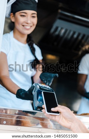 cropped view of woman paying with smartphone near happy asian girl in food truck 
