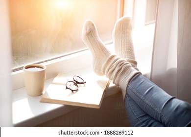 Cropped view of woman putting her feet in warm comfy socks on windowsill with book and coffee, closeup. Unrecognizable lady spending cold day at home, enjoying cozy atmosphere - Powered by Shutterstock