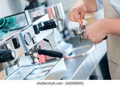 Cropped view of young adult barista in apron uniform working in restaurant, cleaning filter in professional coffee machine, using napkin - Powered by Shutterstock