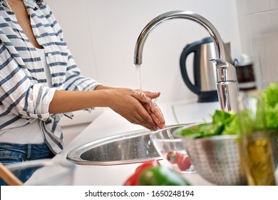 Cropped view of young adult woman standing on kitchen, washing hands in sink before cooking healthy food, holding palms under water