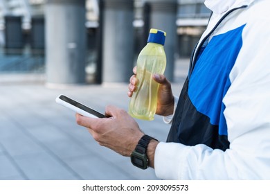 Cropped view of young athlete holding bottle with water, resting after sport training outdoors. Man using online app on modern smartphone, texting a message, having a break after workout
