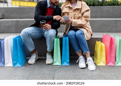 Cropped View Of Young Diverse Couple With Gift Bags And Smartphone Sitting On Stairs, Shopping Online Outdoors. Unrecognizable Woman And Man With Mobile Device Ordering Goods On Web