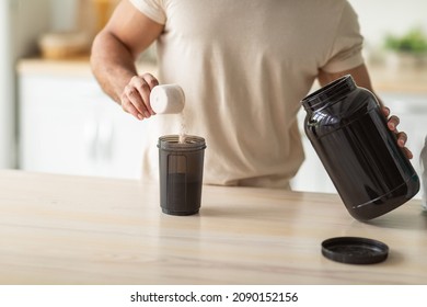 Cropped view of young fit guy making protein shake in kitchen, closeup. Unrecognizable millennial bodybuilder preparing cocktail, using sports nutrition supplement at table indoors