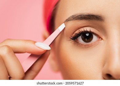 cropped view of young woman with dyed hair holding tweezers while shaping eyebrow isolated on pink