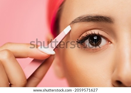 cropped view of young woman with dyed hair holding tweezers while shaping eyebrow isolated on pink
