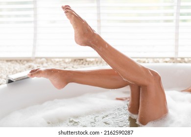 Cropped view of young woman lying in foamy bath, demonstrating silky skin on her legs, enjoying spa treatment at home. Unrecognizable millennil lady relaxing in bathtub at hotel. Domestic depilation