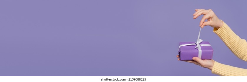 Cropped View Of Young Woman Pulling Ribbon On Wrapped Gift Box Isolated On Purple, Banner