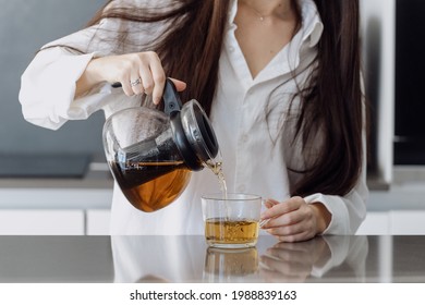 Cropped view of young woman spending morning standing on kitchen, pouring traditional tea from transparent teapot into a glass cup. Female make hot weight loss drink with organic herbs
