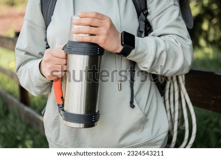 Cropped view of young woman tourist with backpack and fitness tracker holding thermos while hiking with blurred nature at background, trekking through rugged terrain, summer