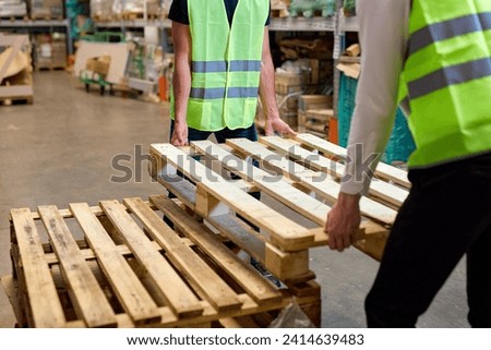 cropped warehouse men hold cargo pallet together with co-worker in workplace area. hardworking strong guys in green vests working uniforms engaged in work in cargo distribution storage