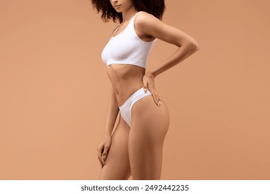 Cropped of woman with curly hair poses in white underwear against a tan backdrop, with her left hand on her hip and her right hand resting on her thigh. - Powered by Shutterstock