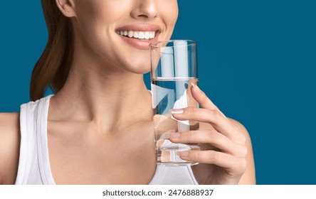 Cropped of woman, dressed in a white tank top, smiling and drinking water from a glass. The background is a solid blue. The image focuses on the womans face, neck, and hand holding the glass - Powered by Shutterstock