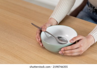 Cropped Of Woman Sitting At Wooden Kitchen Table, Holding Hands On Empty Plate Bowl With Spoon In It, Unrecognizable Poor Hungry Starving Lady Asking For Food, Panorama With Copy Space