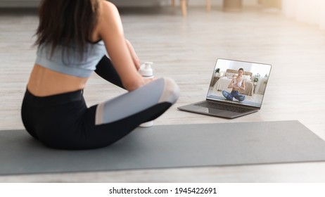 Cropped Of Woman In Sportswear Sitting On Fitness Mat And Looking At Computer Screen At Home, Having Video Call With Yoga Trainer, Using Laptop. Back View Of Sporty Lady Watching Yoga Class Online