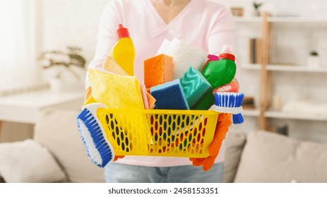 Cropped of woman stands in a sunlit living room, holding a yellow basket filled with various cleaning products and supplies. She is prepared for a housekeeping routine - Powered by Shutterstock