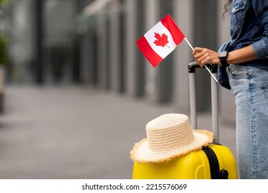 Cropped of woman traveller holding yellow luggage with straw hat on and flag of Canada, going vacation overseas, airport background, copy space. Immigration, education abroad concept
