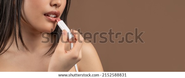 Cropped of young\
asian lady putting lipstick on her beautiful lips, woman applying\
makeup or protecting lips against wind and cold, panorama with copy\
space, brown studio\
background