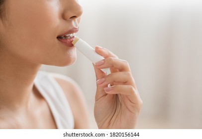 Cropped of young asian woman using lipbalm, bedroom interior, empty space. Side view of unrecognizable lady taking care of her beautiful lips, putting hygienic lipstick at home, closeup