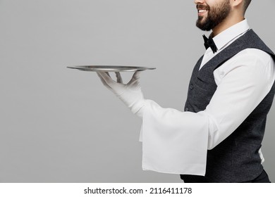 Cropped young barista male waiter butler man wearing white shirt vest elegant uniform work at cafe hold in hand carrying metal tray plate isolated on plain grey background. Restaurant employee concept