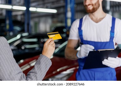 Cropped Young Car Mechanic Man In Blue Overalls Gloves Hold Clipboard, Papers Document Talk With Female Owner Driver Pay Without Cash Work In Vehicle Repair Shop Workshop Indoors. Focus On Credit Card