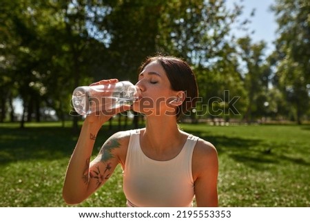 Cropped of young caucasian sportswoman drinking water after training on green lawn. Girl with tattoos and closed eyes wearing sportswear and earphones in sunny park. Healthy lifestyle