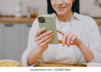 Cropped young happy housewife latin woman wear casual clothes striped shirt hold in hand use mobile cell phone eat breakfast sit at table in light kitchen at home alone. Lifestyle cooking food concept